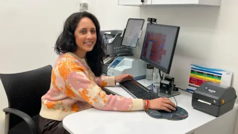 Matt Knight/BBC Hema Patel está sentada en una silla de oficina negra frente a un escritorio blanco. Tiene un monitor de computadora, un teclado y un mouse frente a él y apoya su mano sobre el mouse. Hay varios otros periféricos sobre la mesa. Lleva un suéter estampado de color rosa claro y naranja. Tiene el pelo negro y sonríe a la cámara.