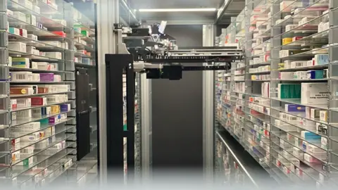 Matt Knight/BBC Un dispositivo robótico negro está colocado en el centro de la habitación. A ambos lados hay estantes con varios tipos de medicamentos. En el otro extremo de la habitación, detrás del robot, hay una puerta gris oscuro.
