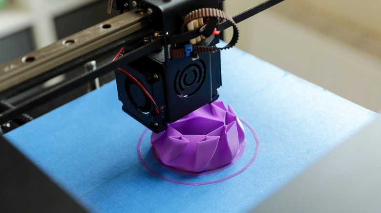 Primer plano de la boquilla de una impresora 3D que imprime un objeto de plástico violeta en una placa de construcción