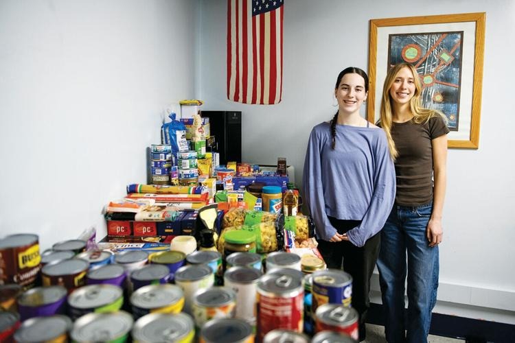 Dos mujeres están de pie junto a una mesa llena de productos enlatados y alimentos envasados en una habitación con una bandera estadounidense y obras de arte enmarcadas en la pared.