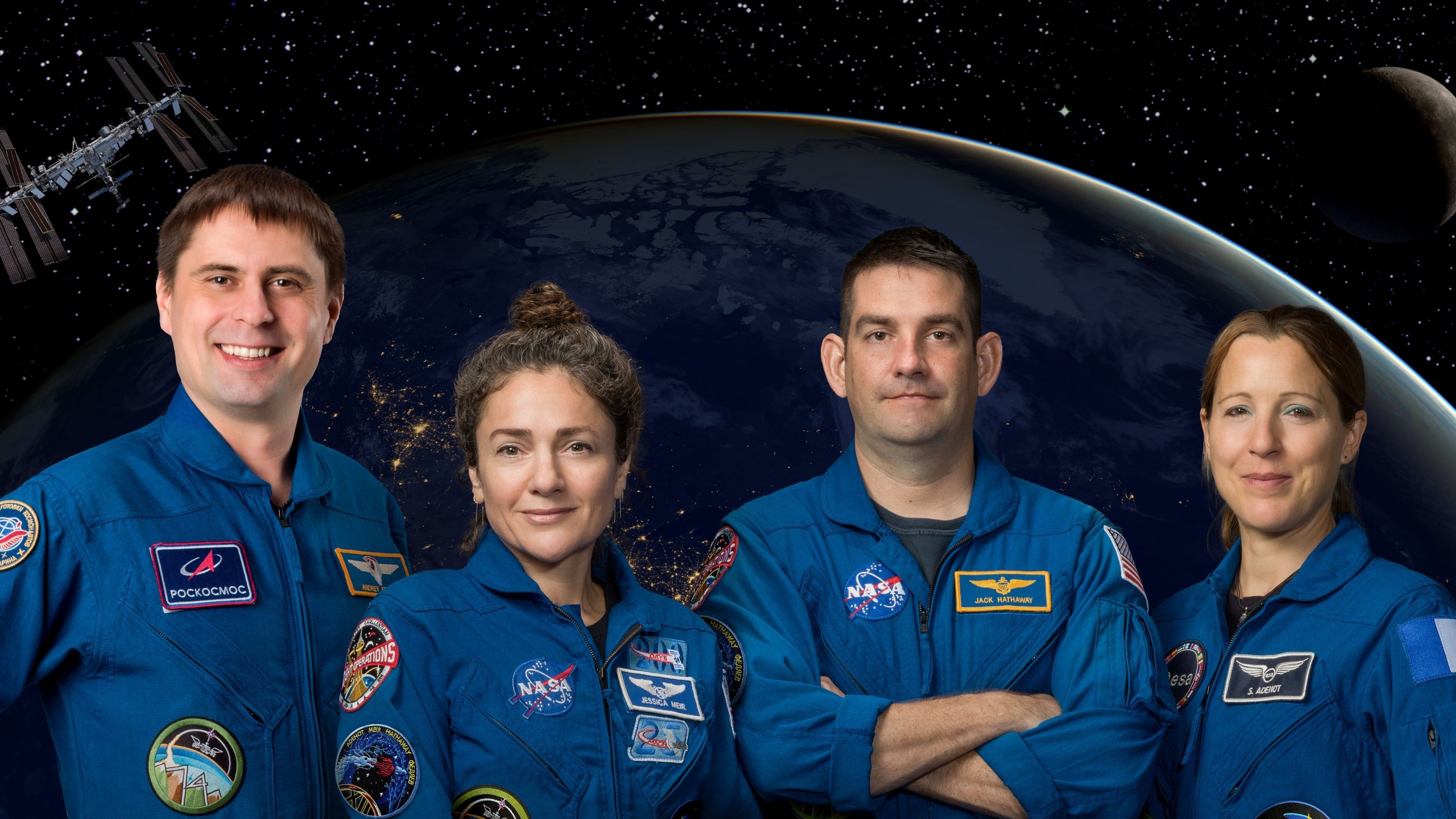 Cuatro personas con trajes azules de pie sobre un fondo de tierra oscura y una luna en sombras, con una pequeña ISS en la esquina superior izquierda.
