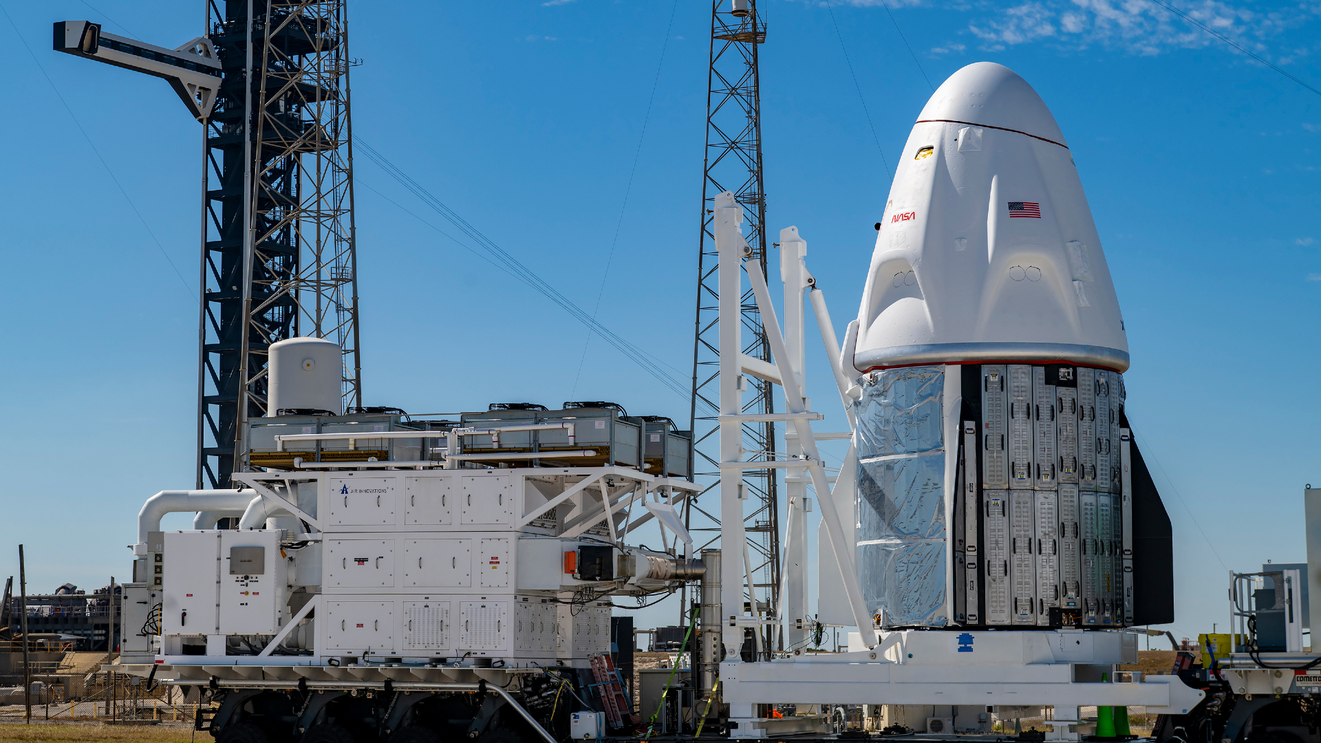 Una nave espacial blanca se encuentra sobre un soporte móvil bajo un cielo azul brillante.